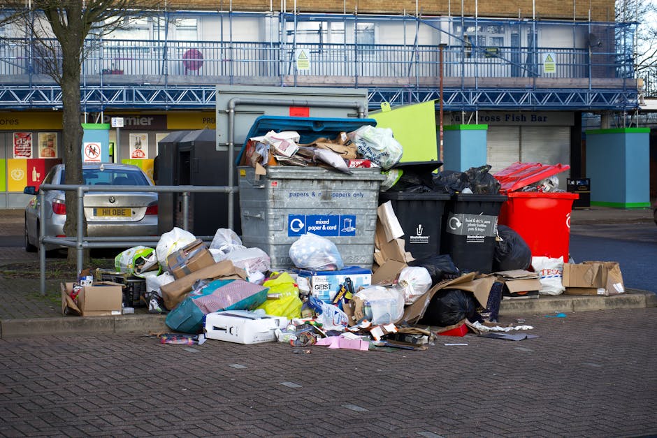 A collection of overflowing rubbish bins and numerous discarded waste items situated on a paved area near a parking lot. The large grey bin, designated for mixed paper and cardboard, is overfilled with crumpled papers, cardboard boxes, and plastic bags spilling onto the ground. Adjacent to it are smaller black bins and a red bin, each filled with varying waste materials such as packaging, plastic wrappers, and other refuse. Surrounding the bins are scattered cardboard boxes, plastic bags, and loose trash, creating a cluttered scene. In the background, a building with a blue scaffolding structure and signage related to food outlets and retail stores is visible, while a parked silver car is partially obscured by a metal railing in the foreground. The environment appears to be an outdoor urban or retail area, illustrating how unmanaged waste can accumulate outside commercial or public premises, emphasizing the importance of efficient rubbish collection and disposal services like those provided by Rubbish Removal Belgravia for private waste handling or alternative collection needs.