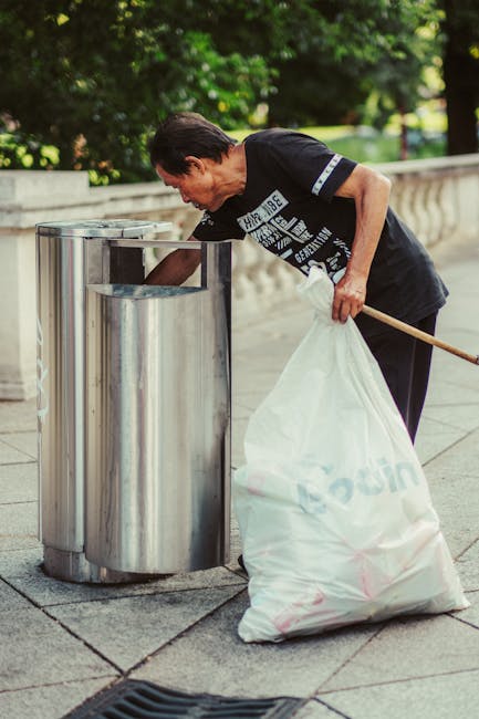 A middle-aged man with dark hair, wearing a black T-shirt with white graphic text and dark trousers, is shown standing on a paved sidewalk in an outdoor park setting, actively disposing of waste into a cylindrical stainless steel rubbish bin with a slot opening. The bin has a smooth, polished finish, reflecting the surrounding environment, and is positioned near a low stone wall with greenery and trees in the background, indicating a peaceful urban park. The man is holding a large white plastic bag filled with rubbish in one hand, with the other hand reaching into the bin to deposit waste. The scene is lit with natural daylight, highlighting the textures of the stainless steel, the fabric of the man's clothing, and the stone paving beneath his feet. This image reflects a typical moment of private waste disposal, which may relate to independent rubbish collection services by companies such as Rubbish Removal Belgravia, facilitating efficient rubbish management outside of local authority systems.