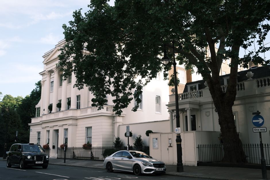 The image depicts a white, multi-story building with classical architectural details such as columns and decorative cornices, situated beside a street. In the foreground, there is a large, mature tree with a thick trunk and dense, dark green foliage that partially obscures the building. Parked along the street in front of the property are a black van and a silver sedan, both aligned parallel to the pavement. The street features a lamppost near the building, and a one-way traffic sign with an arrow, indicating traffic flow directions. The building's exterior appears well-maintained, with a smooth, cream-colored facade and decorative window boxes filled with small plants. The surroundings suggest an urban environment with a neat and orderly appearance, consistent with private property or high-value residential area. This scene may relate to private waste management or rubbish removal services, such as those provided by Rubbish Removal Belgravia, which specialises in local rubbish collection and on-site clearance for well-maintained properties in the area.