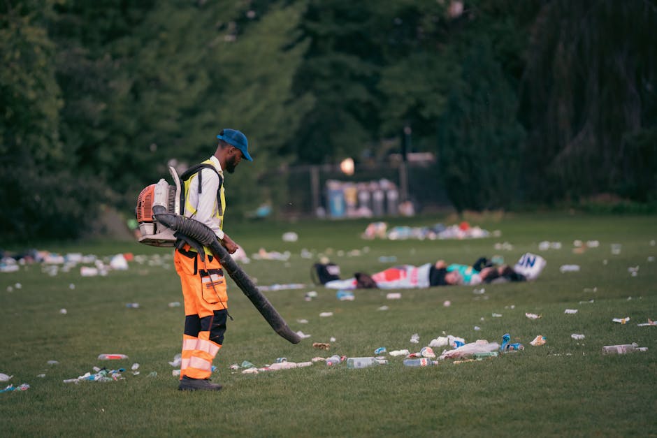 A male waste management worker wearing a blue cap, high-visibility jacket with reflective strips, and orange trousers with black patches is operating a leaf blower or similar equipment on a grassy area, which appears to be an outdoor park or event space. The ground is scattered with various pieces of litter, including plastic bottles, cups, paper debris, and other refuse typical of event waste. In the background, two individuals are lying on the grass, and beyond them, there is a row of trees and a distant utility structure with several composting or waste bins visible, suggesting a managed outdoor environment. The lighting indicates early evening or late afternoon, casting a neutral tone over the scene. The presence of the worker and the scattered rubbish implies an effort in waste collection or site cleanup, aligning with private rubbish removal services such as those provided by Rubbish Removal Belgravia, particularly for clean-up after events or receptions in the area.