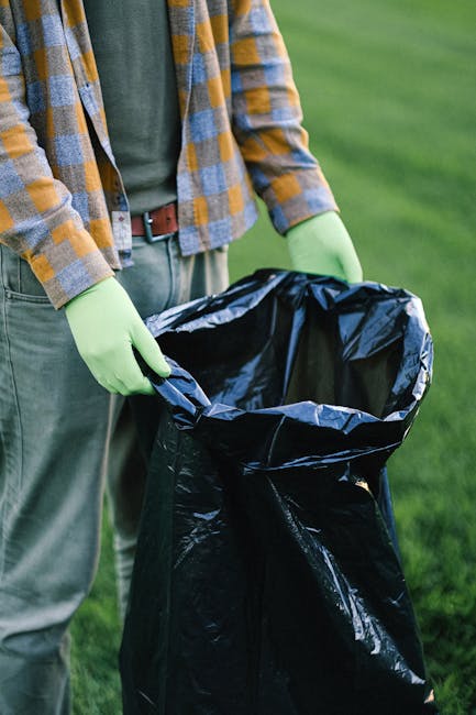 The image shows a person standing outdoors on a grassy area, holding open a large, black trash bag lined with shiny, reflective material. The individual is wearing a plaid shirt with shades of orange, gray, and blue, along with light green gloves for handling the rubbish. Their lower body is dressed in gray pants secured with a brown belt. The background features a well-maintained lawn or garden setting, with vibrant green grass extending into the distance. The scene suggests a rubbish collection or garden waste disposal activity, consistent with private waste handling or on-site clearance services offered by Rubbish Removal Belgravia. The focus on the trash bag and the protective gloves highlights an emphasis on cleanliness and safety during waste removal operations, aligning with themes of alternative rubbish collection methods outside local authority services.