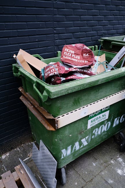A green wheeled skip positioned on a paved surface next to a dark brick wall, filled with bags of building materials such as red Flexible PVC insulation and other packaging, indicating recent waste collection or clearance activity by Rubbish Removal Belgravia. The skip's side features white text and a green label with company contact information, and a wooden plank rests on the top edge, suggesting an on-site clearance or private waste disposal process rather than council take-back. The surrounding environment includes a small section of pavement and some construction debris visible at the bottom left corner, emphasizing the context of rubbish removal services tailored for local property cleanouts or renovation waste streams within an urban setting.