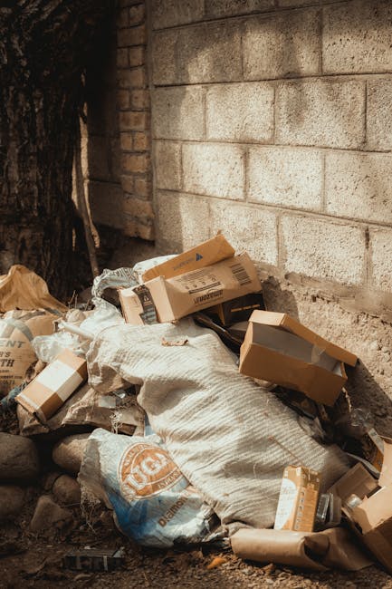 A pile of various discarded cardboard boxes, some flattened and some partially assembled, is seen against a weathered brick wall and a concrete block wall in an outdoor space. The boxes are of different sizes, with some featuring printed labels and barcodes, and are interspersed with a large, textured fabric sack that appears filled and closed at the top. Surrounding the trash are small rocks and loose debris, with a dark tree trunk visible to the left side. The scene suggests an area designated for discreet rubbish removal or private waste disposal, with the materials and environment indicative of a clearing spot for scheduled rubbish collection services, such as those provided by Rubbish Removal Belgravia. The lighting is natural, creating soft shadows that highlight the textures and materials, emphasizing the organized yet temporary nature of this waste pile in an urban setting. This scene exemplifies typical outside rubbish accumulation awaiting professional clearance, aligning with the services of independent waste handling and rubbish removal specialists specialized in discreet, on-site clearance within residential or commercial properties.