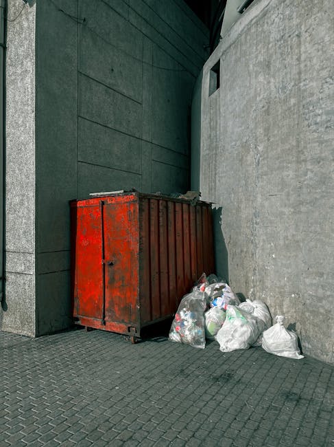 A green wheeled skip positioned on a paved surface next to a dark brick wall, filled with bags of building materials such as red Flexible PVC insulation and other packaging, indicating recent waste collection or clearance activity by Rubbish Removal Belgravia. The skip's side features white text and a green label with company contact information, and a wooden plank rests on the top edge, suggesting an on-site clearance or private waste disposal process rather than council take-back. The surrounding environment includes a small section of pavement and some construction debris visible at the bottom left corner, emphasizing the context of rubbish removal services tailored for local property cleanouts or renovation waste streams within an urban setting.