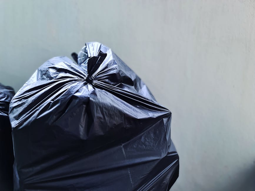 Close-up image of several large black plastic rubbish bags tightly tied at the top, positioned against a plain light-colored wall. The bags are made of shiny, smooth plastic material with visible creases and folds, some reflecting light. The bags occupy the foreground of the image, with the top portion prominently displayed, showing a twisted knot and gathered plastic. In the background, part of another similar bag is partially visible, suggesting they are stored together in an indoor or sheltered area, possibly prepared for collection or disposal. The neutral lighting emphasizes the glossy surface of the bags without any shadows or environmental details. This scene relates to private waste handling and rubbish removal services, such as those provided by Rubbish Removal Belgravia, especially in contexts requiring alternative collection methods for bulky or excess waste.